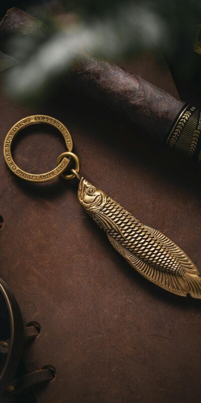 Close-up of a brass fish keychain elegantly resting on a leather journal, accompanied by a vintage cigar.